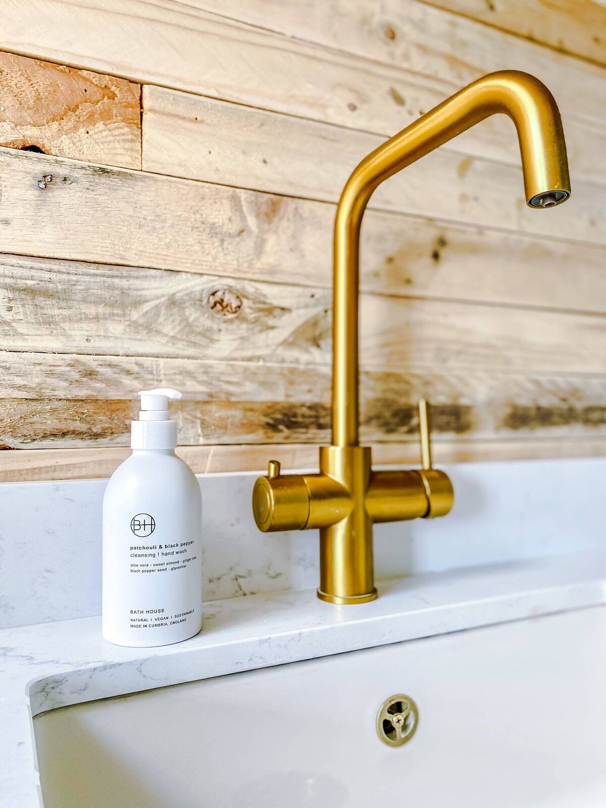A bathroom with a white sink, a gold faucet, and a bottle of bathhouse cleansing hand wash.
