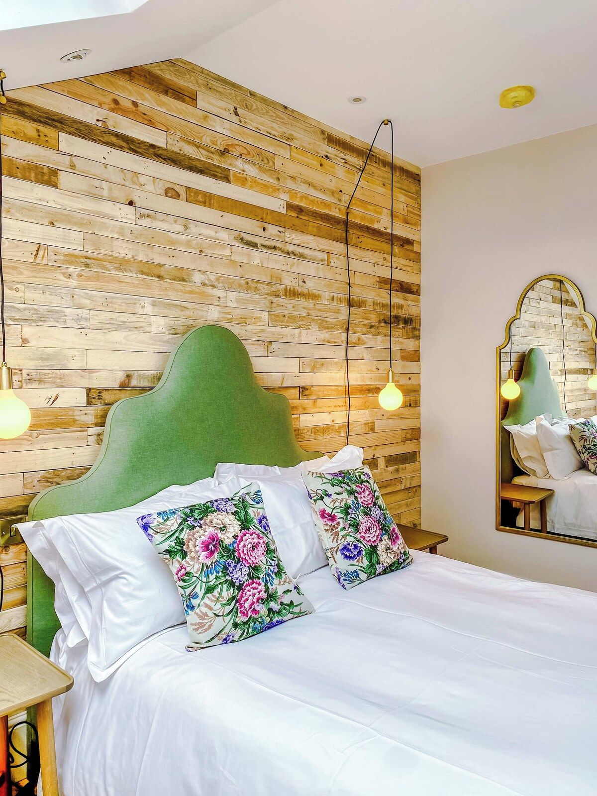 A bed with a green headboard, white bedding, and floral pillows. A wooden wall with a herringbone pattern and a mirror with a gold frame are visible.