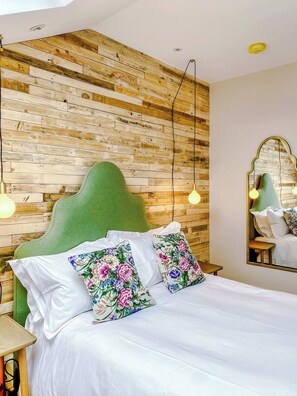 A bed with a green headboard, white bedding, and floral pillows. A wooden wall with a herringbone pattern and a mirror with a gold frame are visible. - Railway House -Platform 8, 2 Bedroom Apartment. (York)