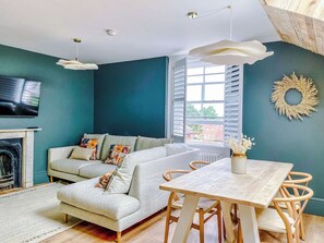 A living room with a sofa, a dining table, and chairs. There is a fireplace, a television, and a window with blinds.