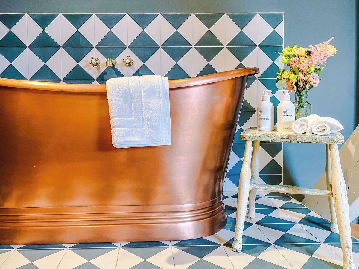 Une salle de bain avec une baignoire en cuivre, un petit tabouret sur lequel sont posés des articles de toilette et un vase rempli de fleurs.