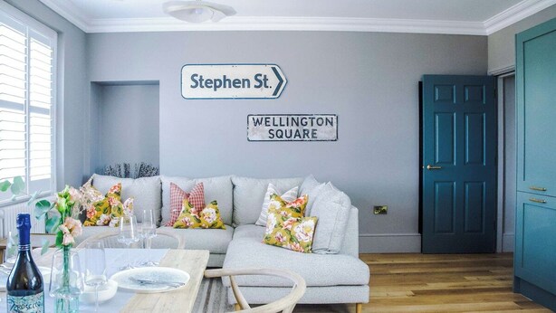 Quarto com sofá cinzento, mesa de jantar posta com pratos e copos, uma garrafa de Prosecco e uma placa na parede indicando 'Stephen St.' e 'Wellington Square'.