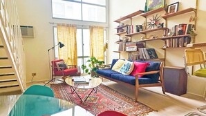 Uma sala de estar ampla com uma janela grande, um sofá vermelho, um sofá azul, uma mesa de centro e uma estante repleta de livros e objetos decorativos.