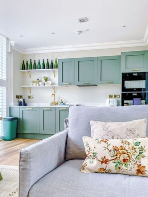 Una cocina moderna con armarios azul verdoso, un sofá gris con cojines estampados y un cubo de basura a la vista.