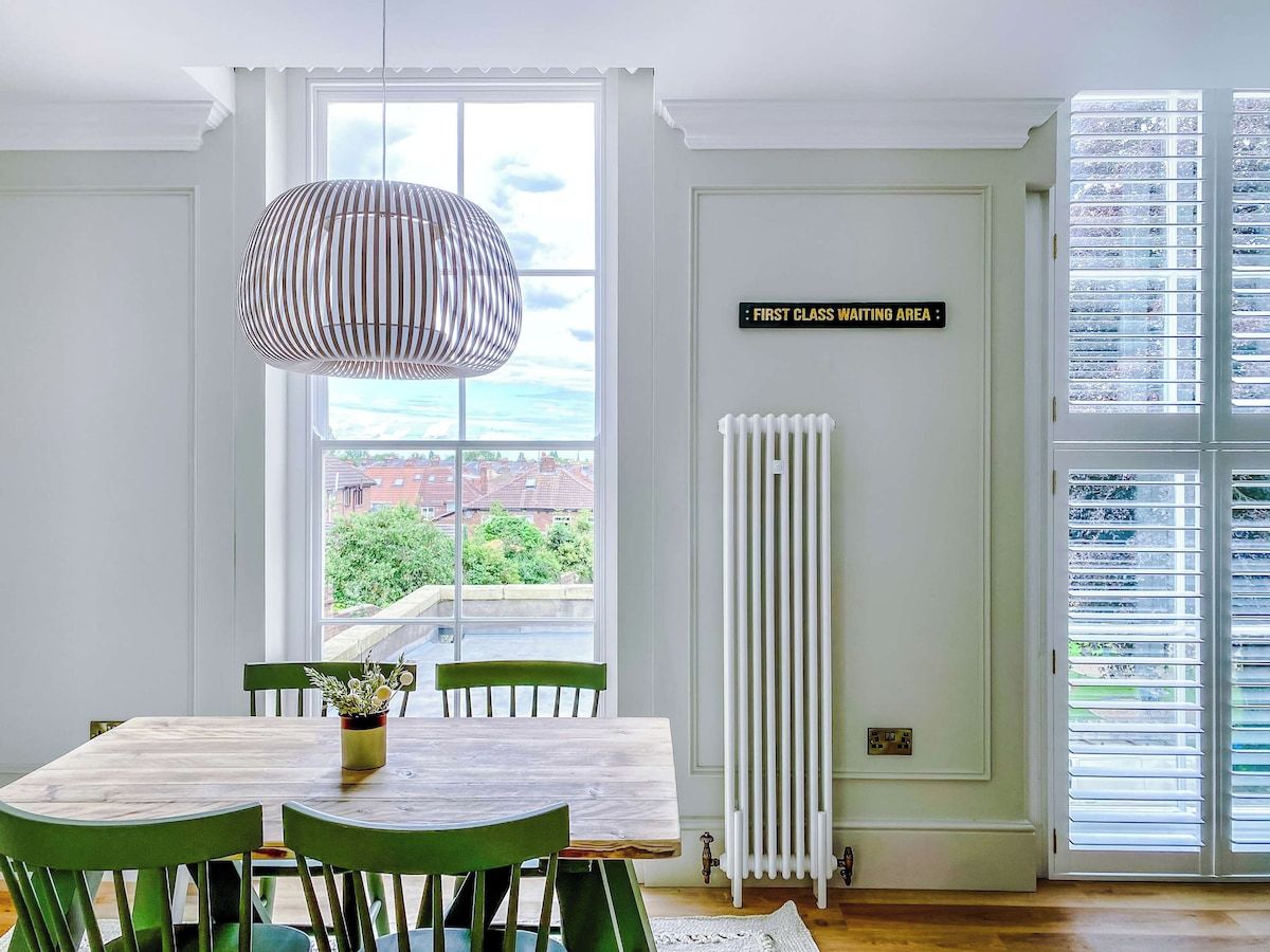 A dining area with a table, chairs, a hanging light fixture, a view through a window, and a sign indicating a first-class waiting area.