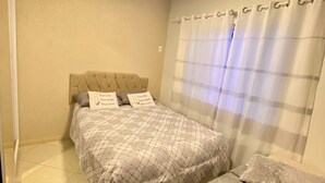 A bedroom with two beds, beige walls, a ceiling light, and a window with curtains.