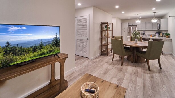 A modern kitchen with white cabinets, a dining area with a wooden table and chairs, and a television displaying a scenic view. - Ruby 314: 2BD | Great LOCATION + Shared Amenities! (Pigeon Forge)