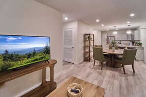 A modern kitchen with white cabinets, a dining area with a wooden table and chairs, and a television displaying a scenic view.