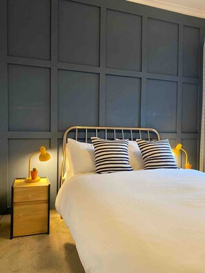 Een opgemaakt bed met hoofdeinde, twee bedlampen en een klein houten nachtkastje met een lamp.