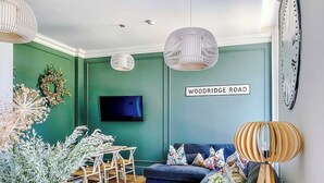 Uma sala de estar com parede verde, televisão de tela plana, sofá azul, luminária de chão de madeira e um letreiro com a inscrição 'WOODRIDGE ROAD'.