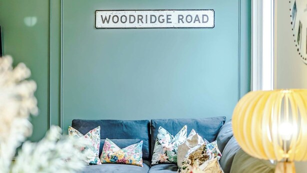 Quarto com sofá azul, luminária pendente e placa na parede com a inscrição 'WOODRIDGE ROAD'.