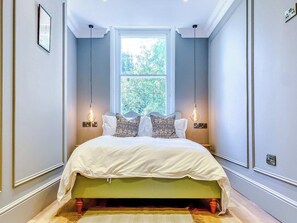 A bedroom with a large bed, two bedside lamps, a window with a view of greenery, and a light switch on the wall.