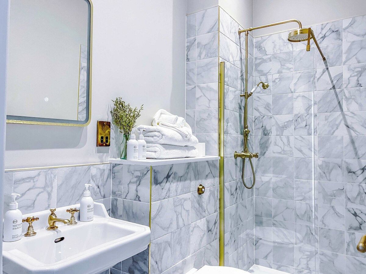 A bathroom with a marble shower, a sink with gold fixtures, and a mirror.