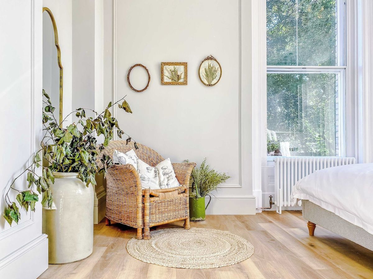 A room with a wicker chair, a large vase with greenery, a round rug, and a bed with white bedding.
