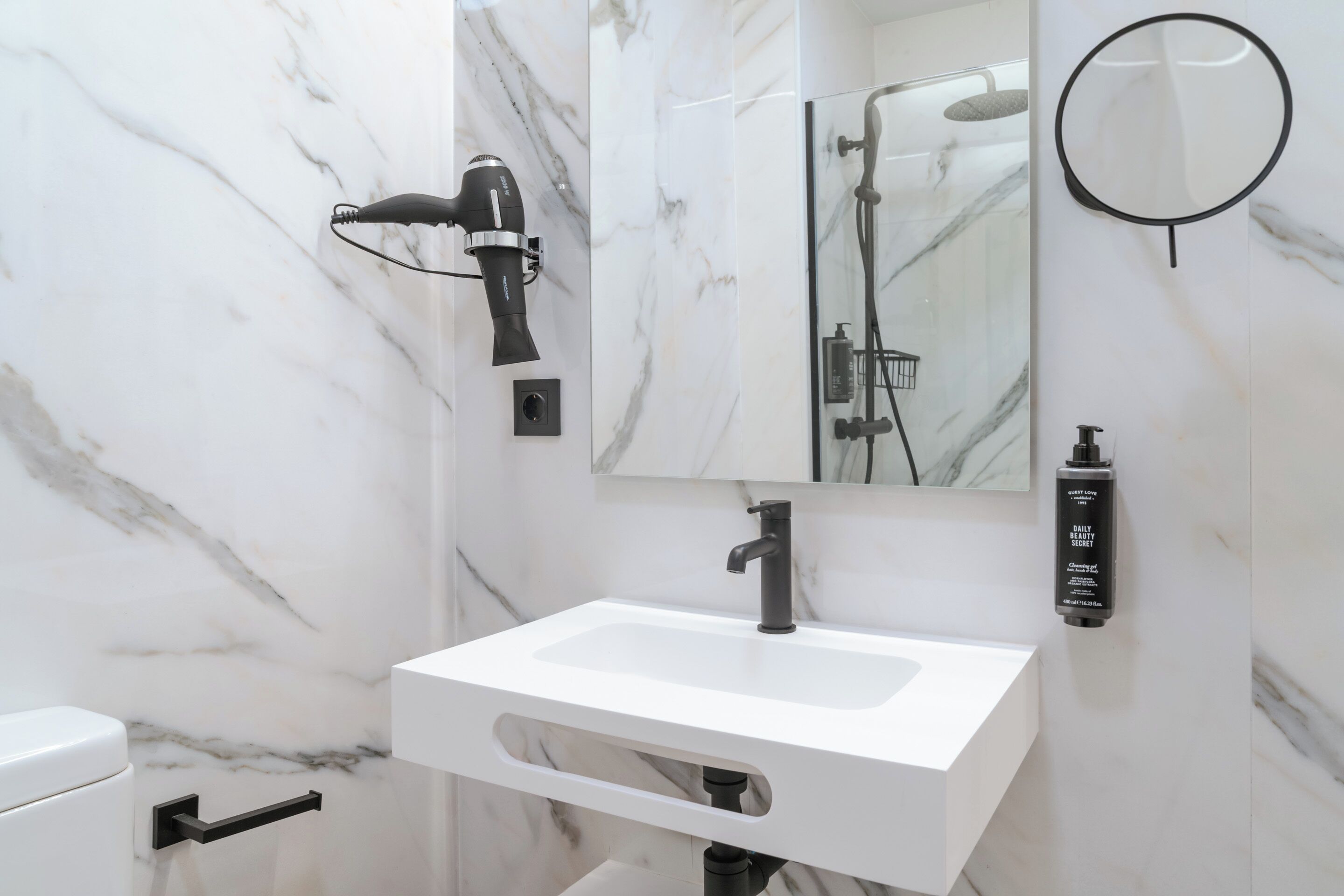 Un baño con lavamanos blanco, secador de pelo de mano, espejo de mano y una botella de jabón.