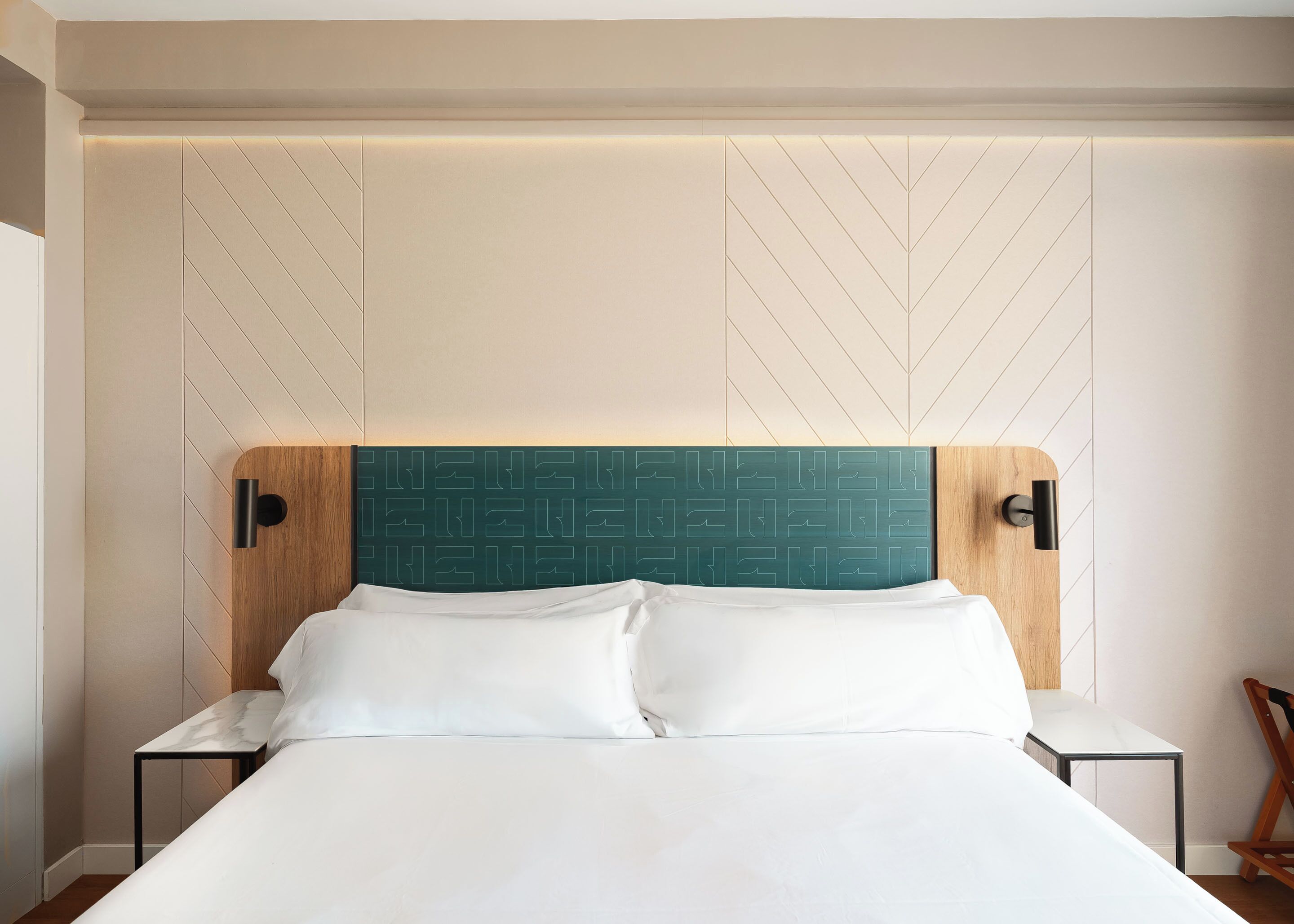 Una habitación de hotel moderna con una cama grande, cabecera de madera con patrón geométrico y dos mesitas de noche con lámparas.
