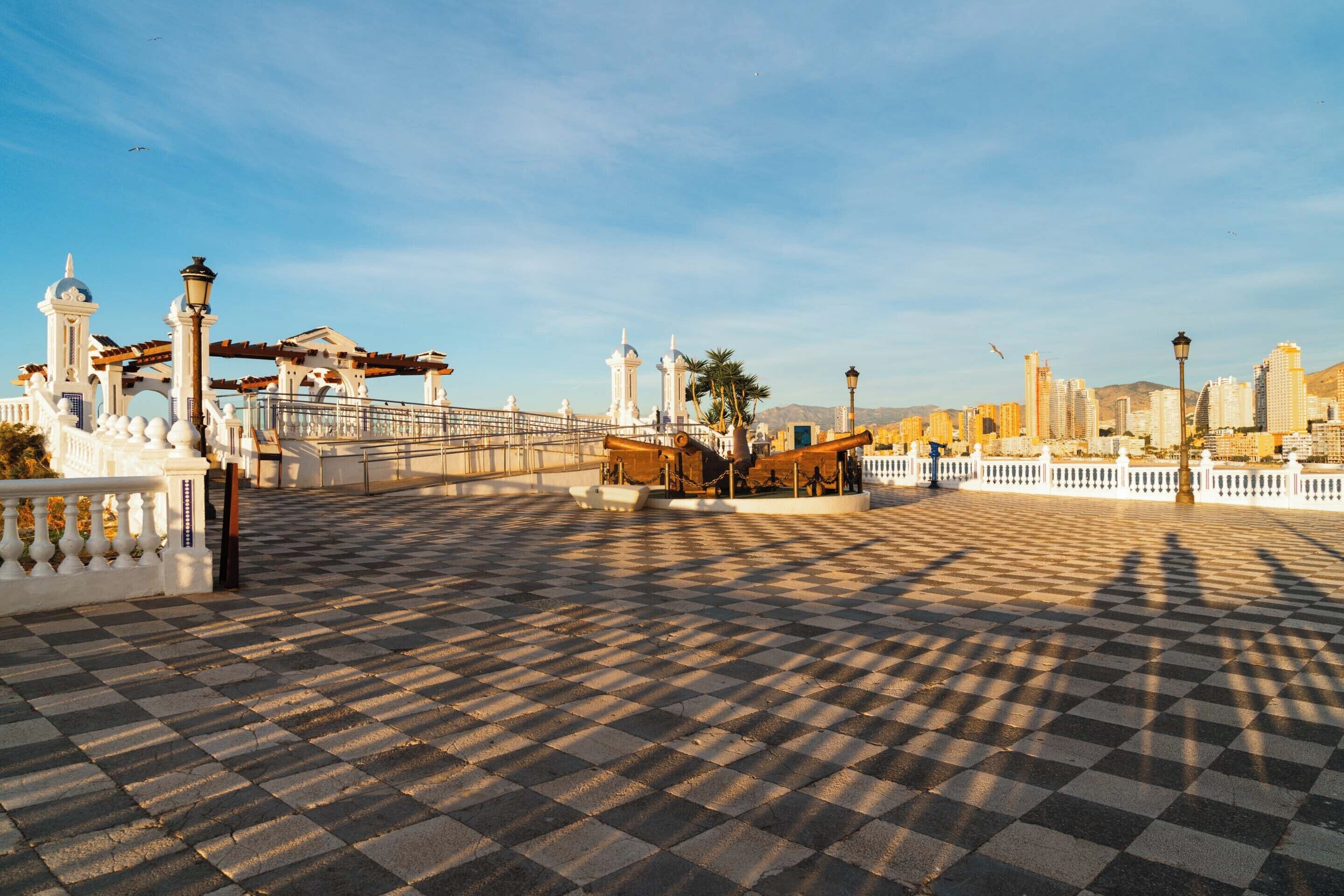 Una plaza a cuadros con un puente, edificios blancos y el perfil de una ciudad al fondo.