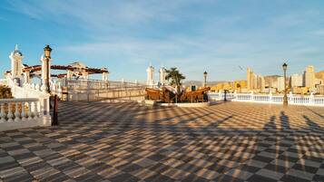 Una plaza a cuadros con un puente, edificios blancos y el perfil de una ciudad al fondo.