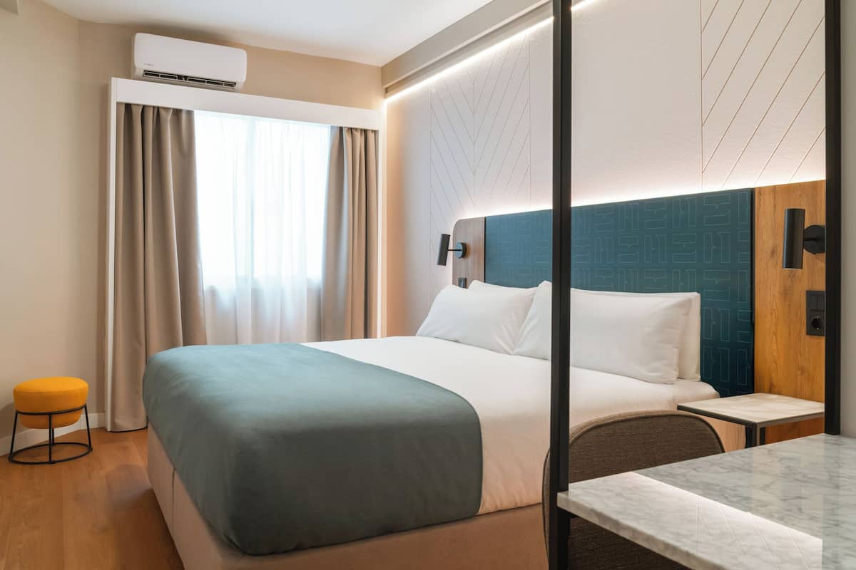 A modern hotel room with a large bed, a small table, a chair, a stool, and an air conditioning unit.