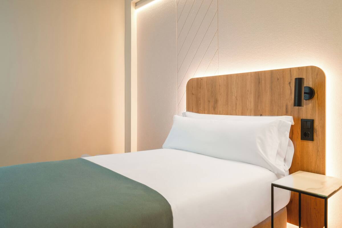 Una cama bien hecha con sábanas blancas y una manta verde, un cabecero de madera con una lámpara de techo y una mesita de noche con una lámpara.