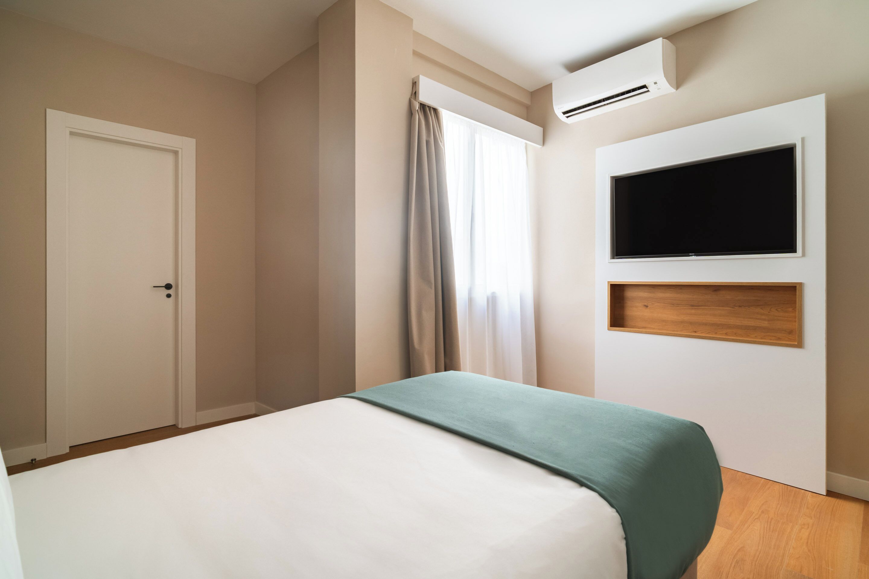 A hotel room with a bed, a TV mounted on the wall, and an air conditioning unit.