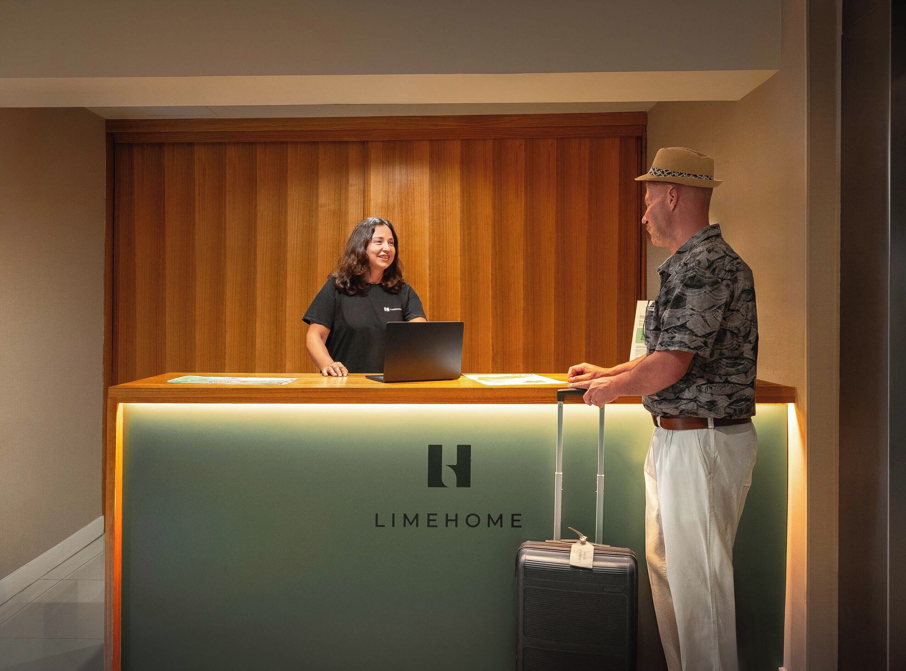 A reception desk with a laptop, a man standing beside it, and a suitcase.