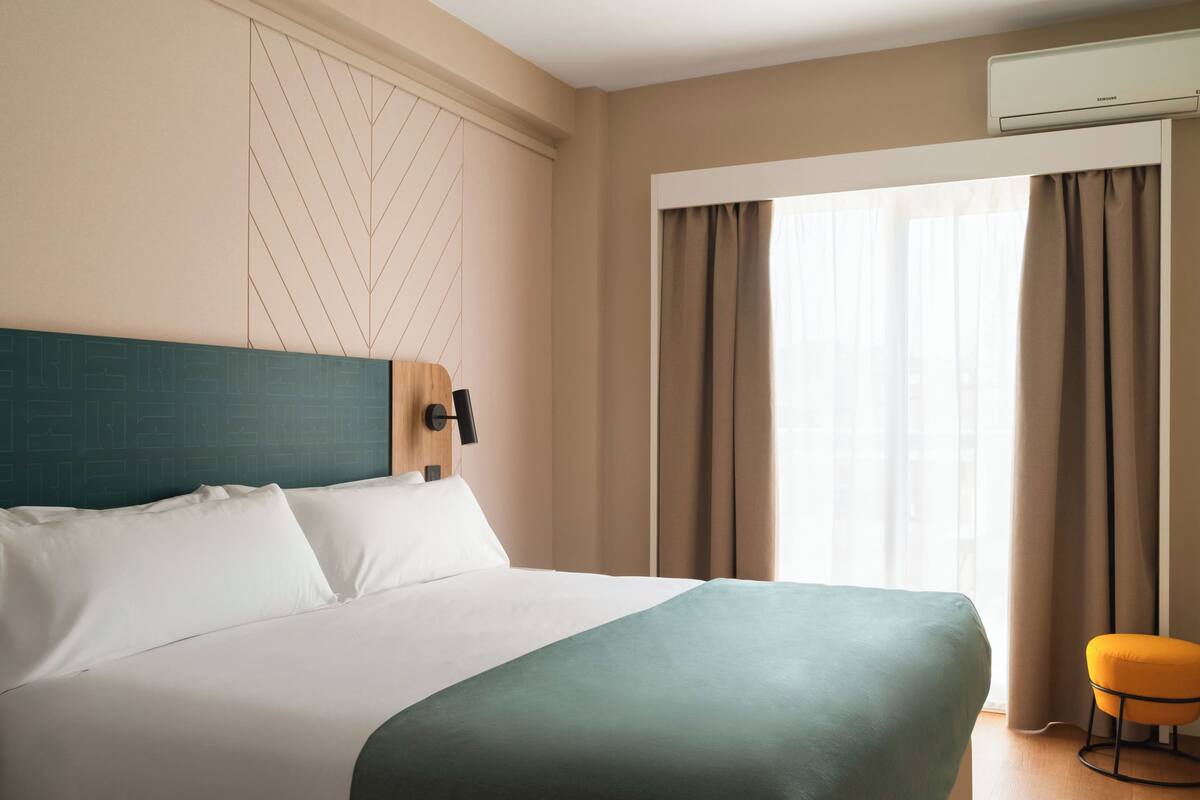 A hotel room with a bed, a wall-mounted air conditioner, a window with curtains, and a small stool.