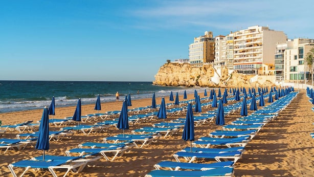 Una playa con hileras de tumbonas y sombrillas azules, un acantilado rocoso y edificios al fondo.