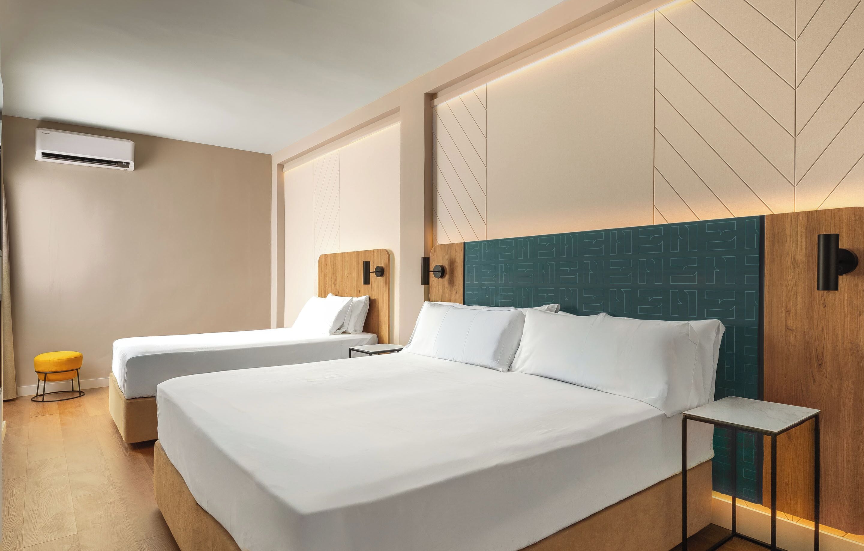 Ein Hotelzimmer mit zwei Betten, einem kleinen Hocker, einer Klimaanlage und einem Nachttisch.