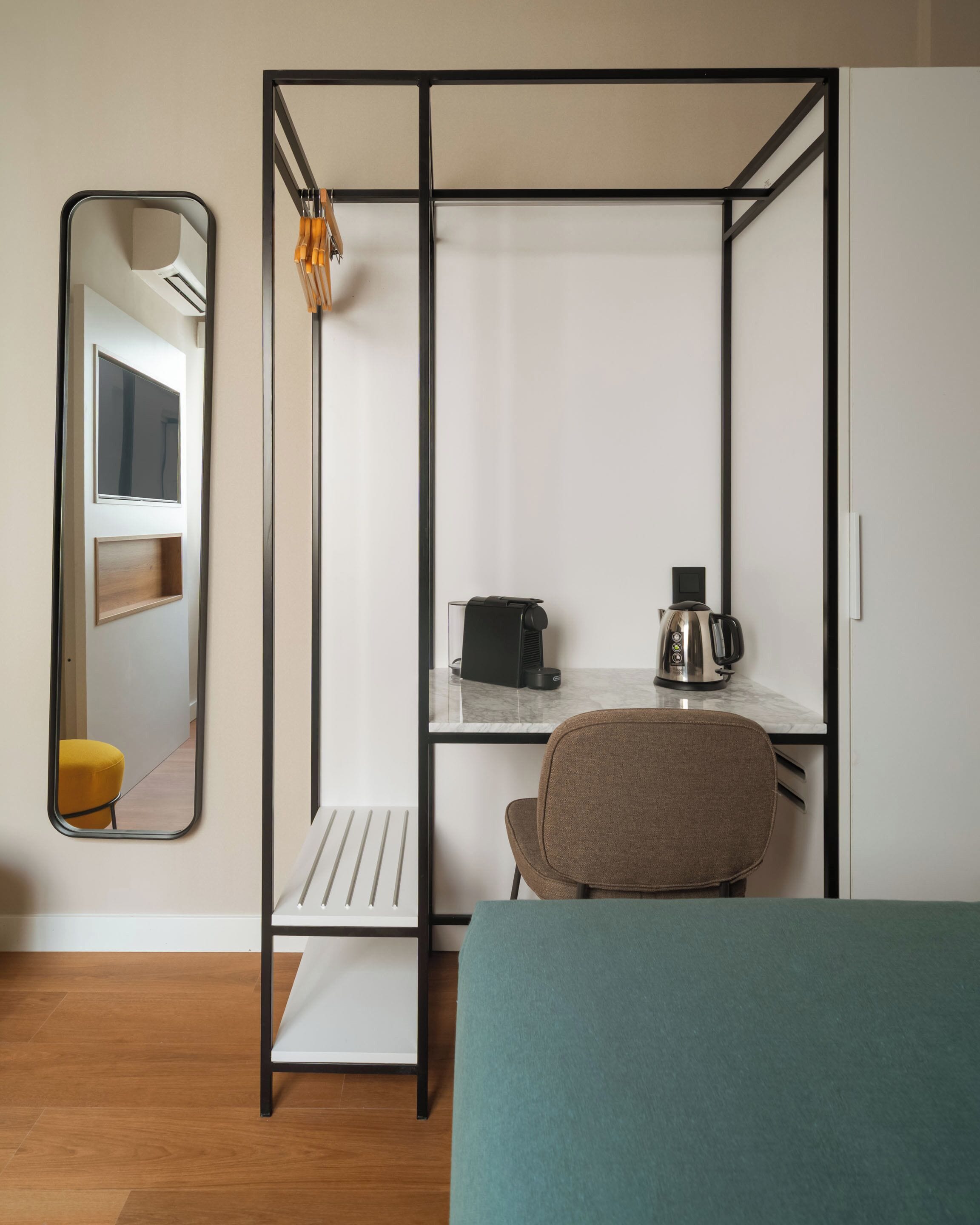 A compact room with a bed, a desk with a chair, a coffee maker, and a mirror.