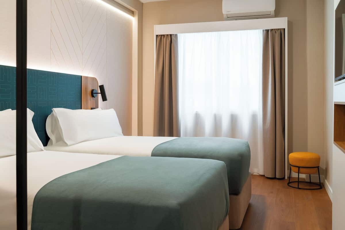 A hotel room with two beds, a window with curtains, an air conditioning unit, and a small stool.