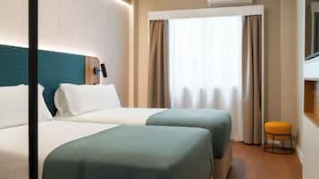 A hotel room with two beds, a window with curtains, an air conditioning unit, and a small stool.