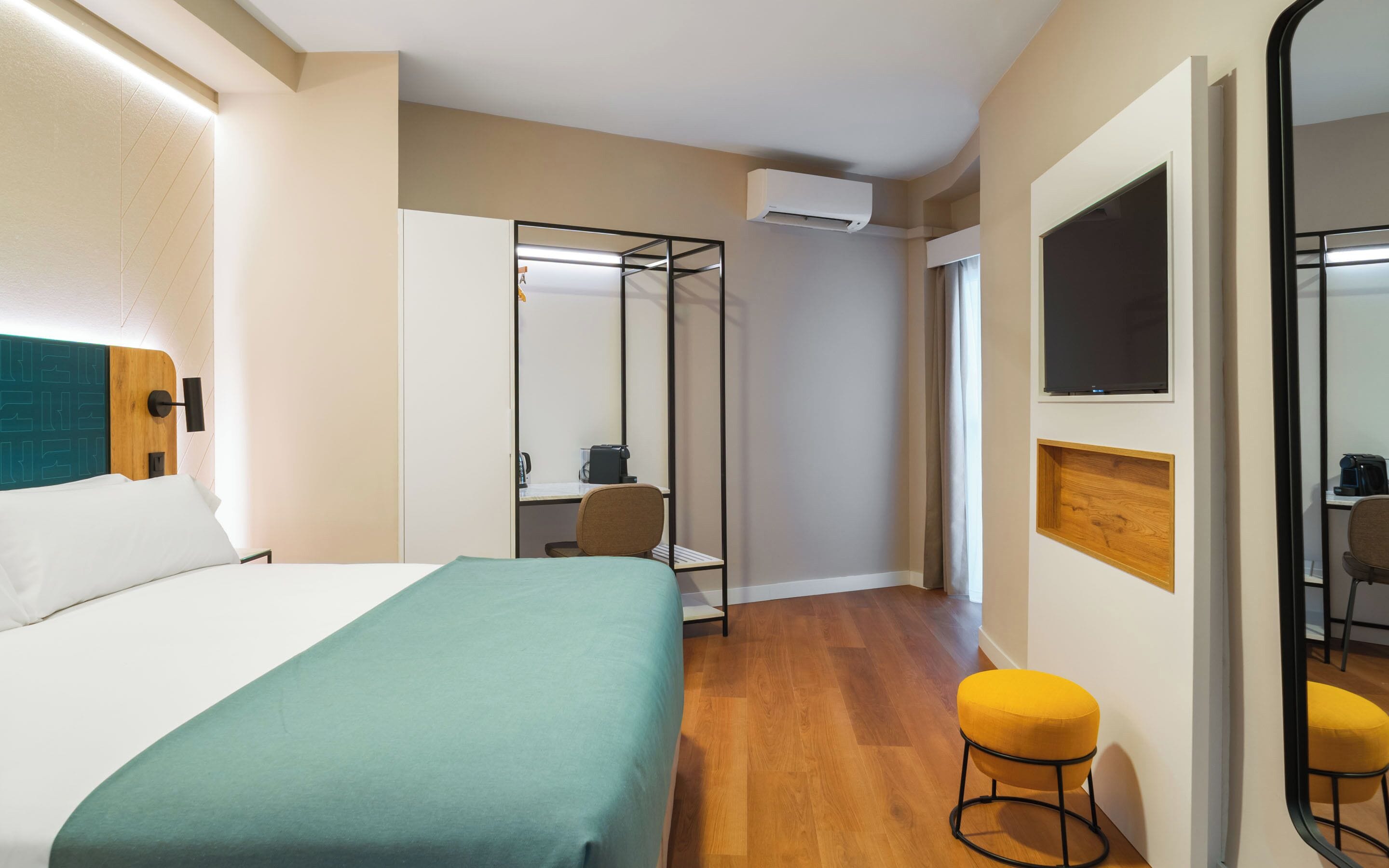 Una habitación de hotel con una cama, un escritorio, una silla, un taburete, un equipo de aire acondicionado y un espejo.