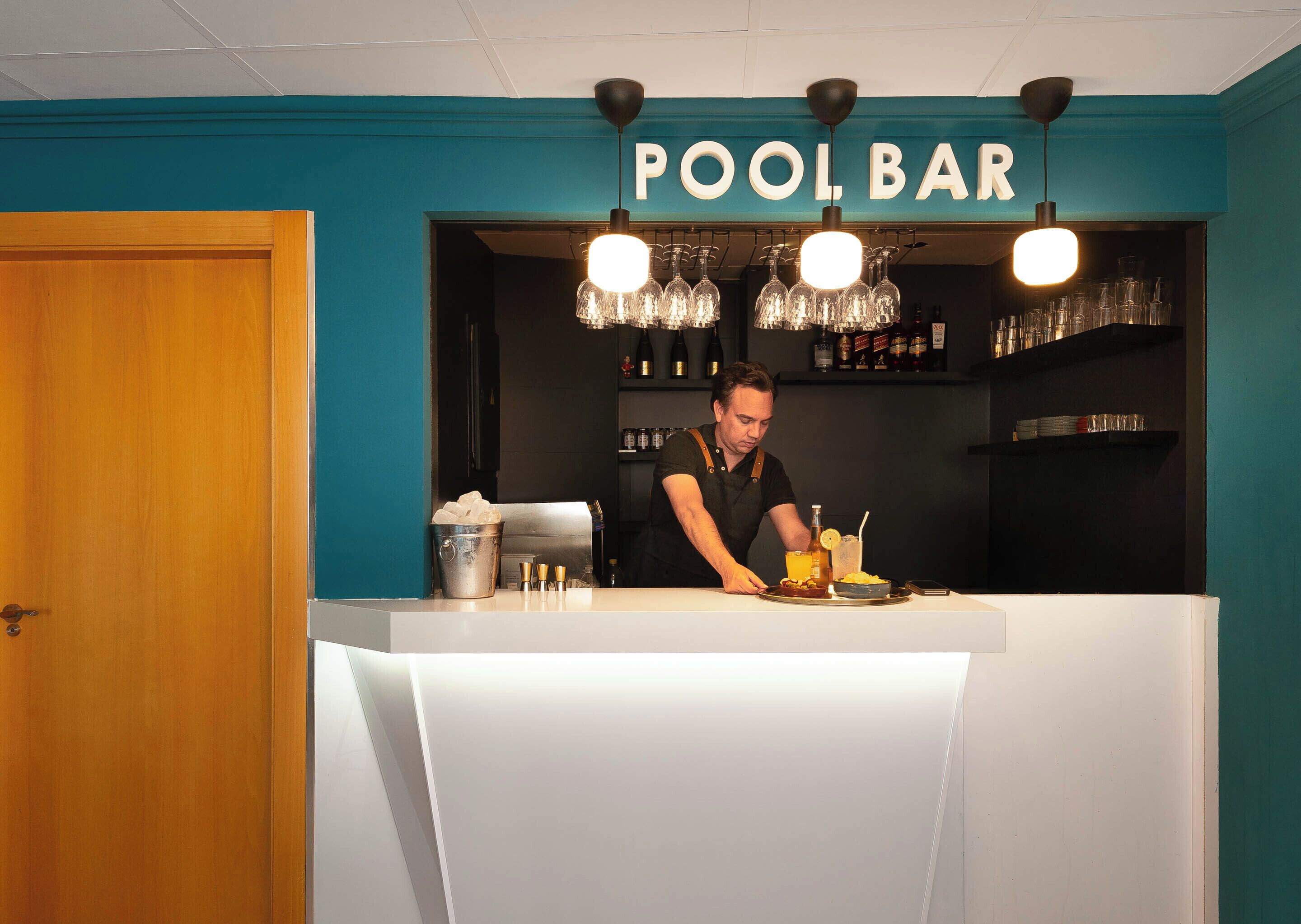 A bartender at a pool bar counter with a teal wall and a sign above.