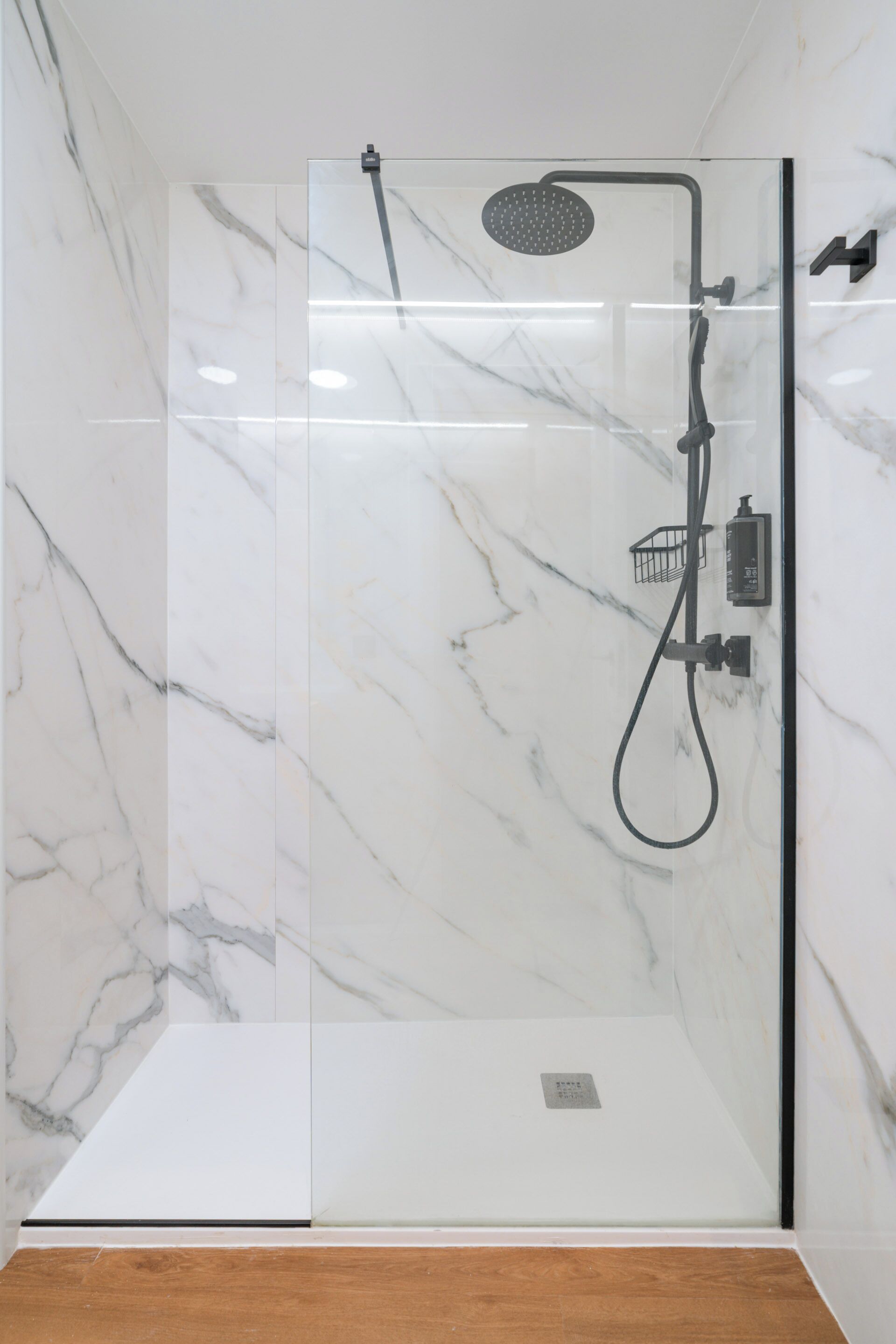 Une douche en verre, dotée d’un pommeau de douche et d’un flexible à main, le tout installé sur des murs en marbre.