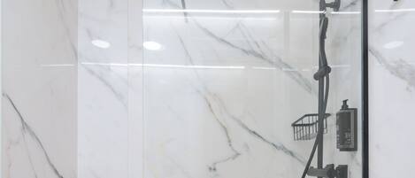 Une douche en verre, dotée d’un pommeau de douche et d’un flexible à main, le tout installé sur des murs en marbre.