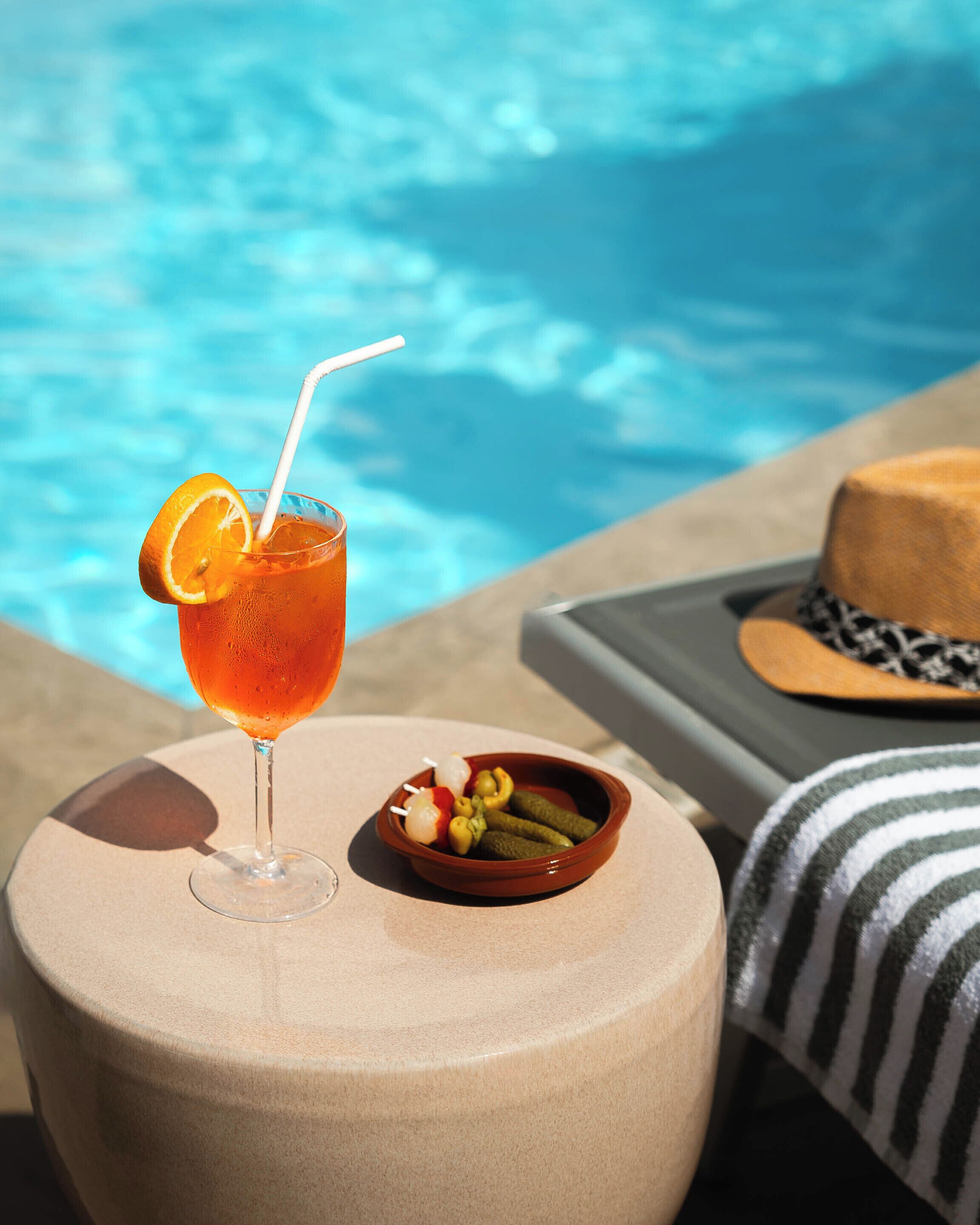 A glass of orange juice with a straw and a slice of orange on a round table by a poolside.