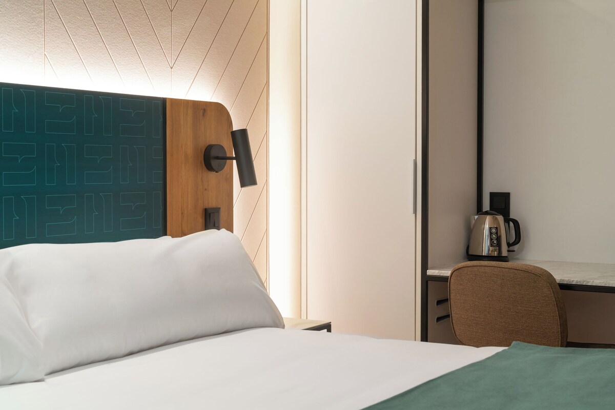 A hotel room with a bed, a desk, and a chair. There is a kettle on the desk.