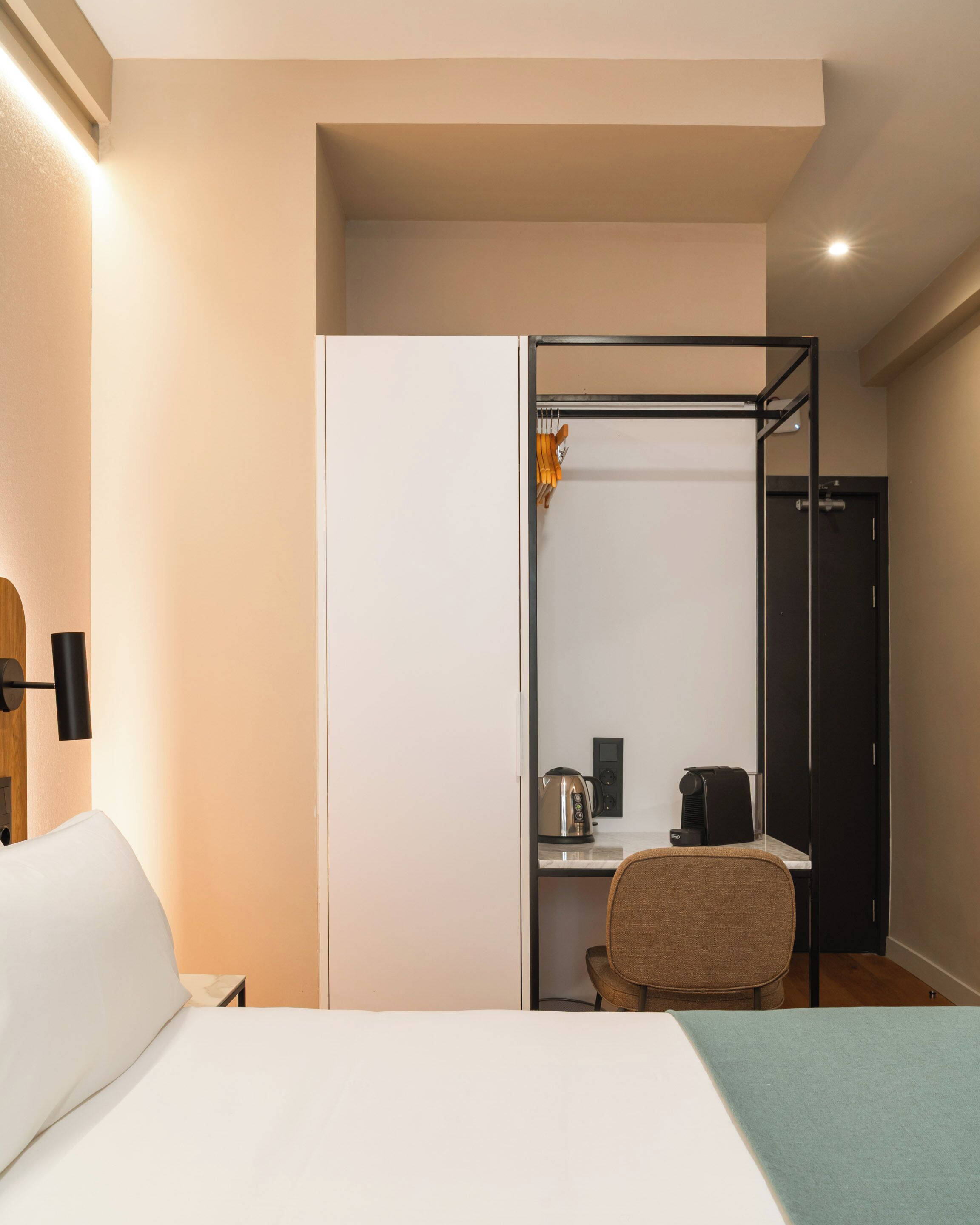 A hotel room with a bed, a desk with a chair, a kitchenette, and a wardrobe.