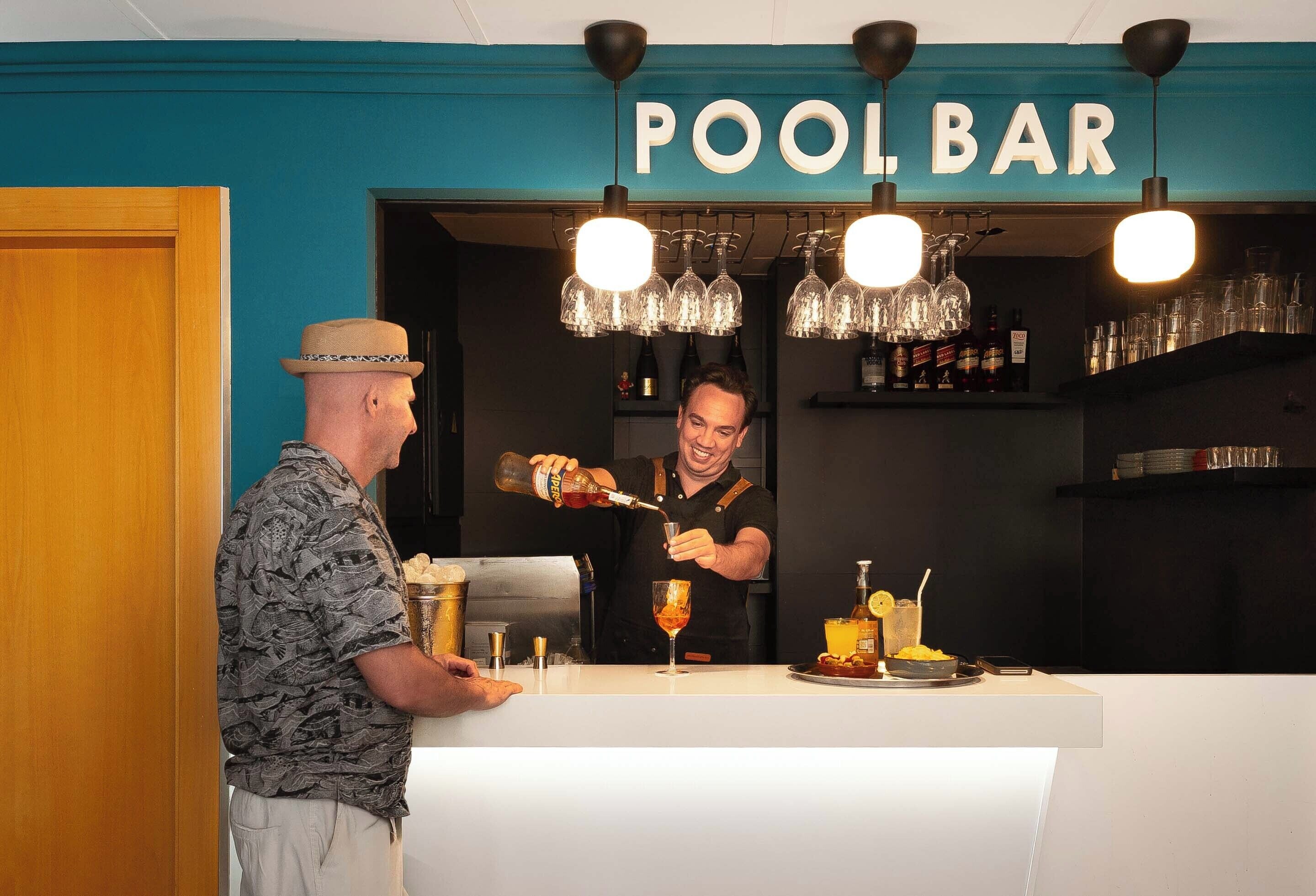 Un espace bar avec une enseigne indiquant « BAR AU BORD DE LA PISCINE » et un barman qui prépare des boissons.