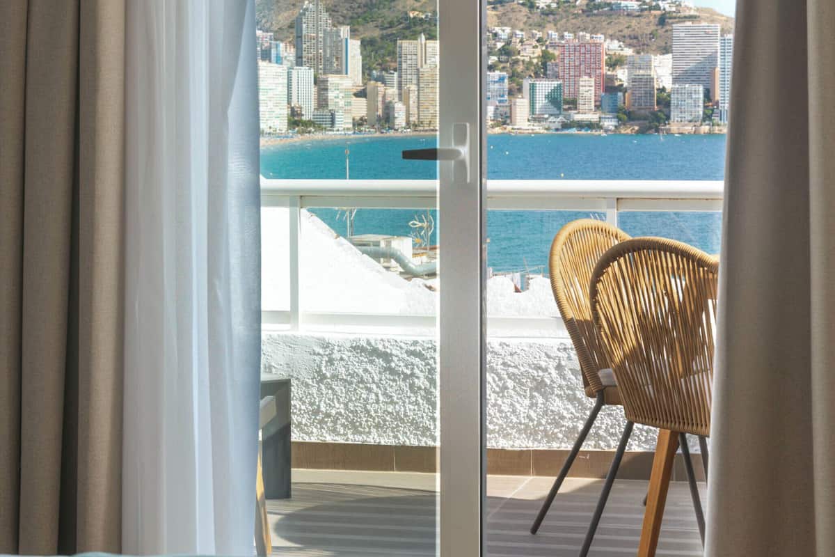 A room with a bed, a chair, and a view of a cityscape and water through a large window.