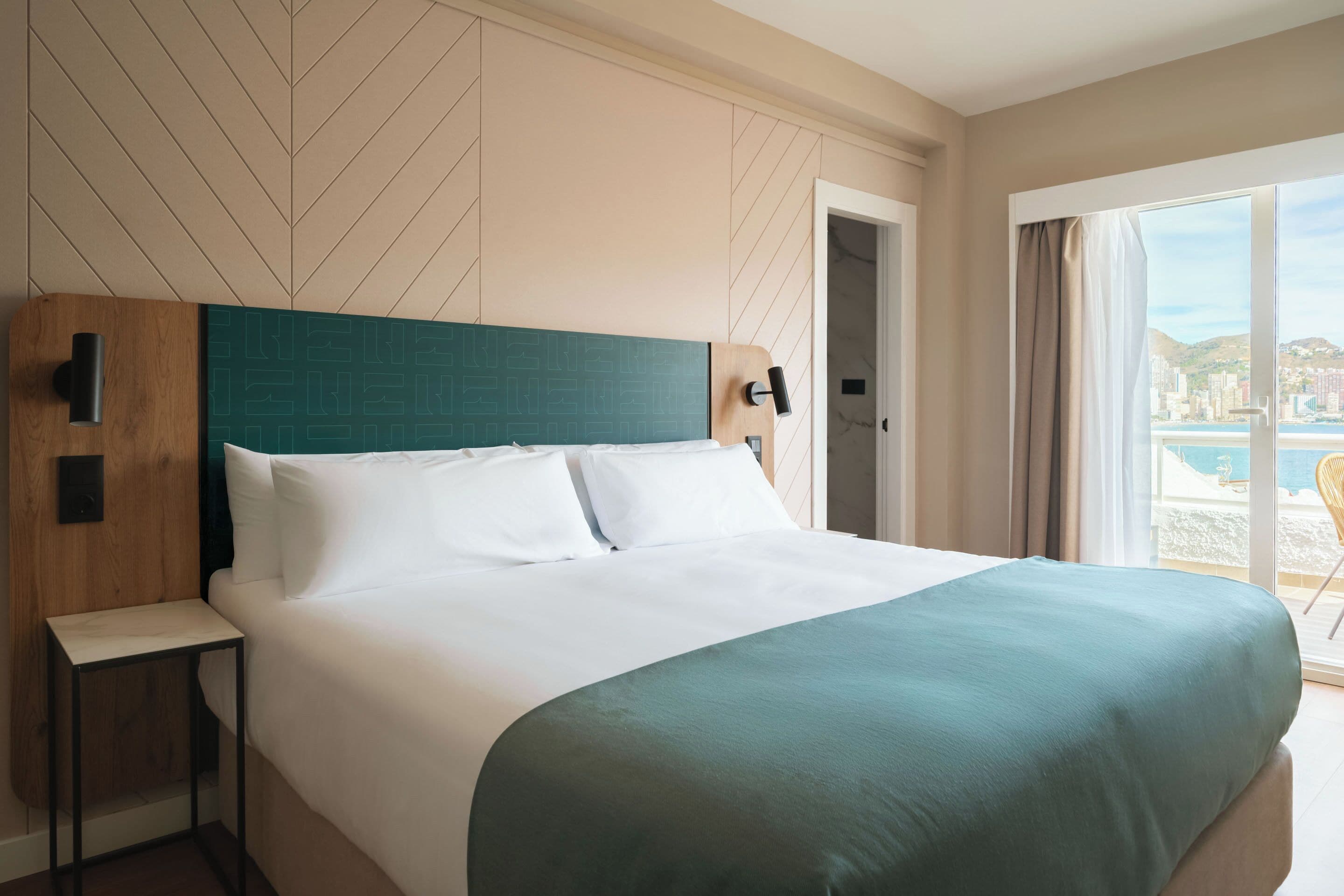 Camera d'albergo con un letto grande, una testiera in legno, un comodino e vista sulla città attraverso un'ampia finestra.