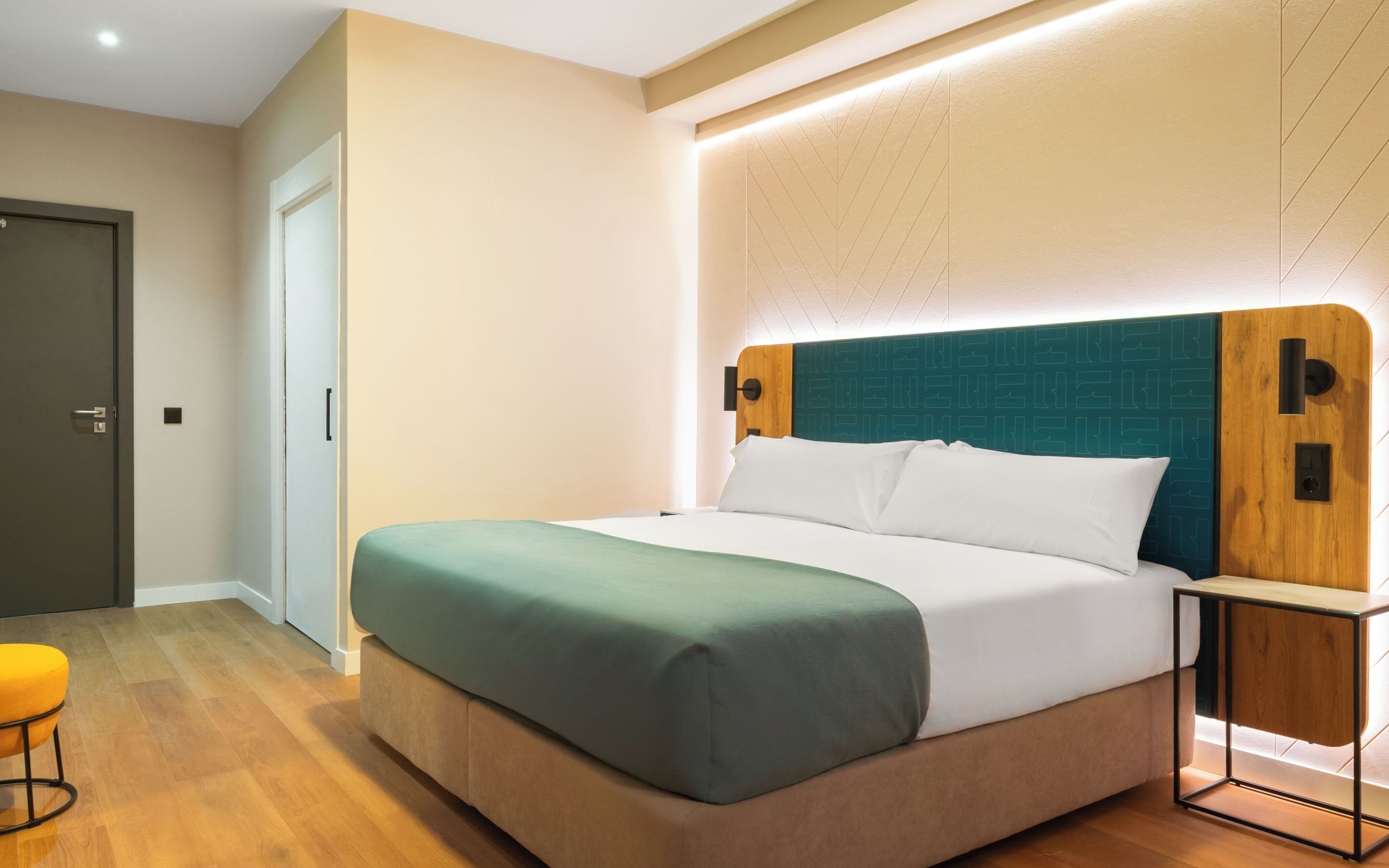 Una habitación de hotel moderna con una cama grande, un cabecero de madera, una mesita de noche y un taburete pequeño naranja.