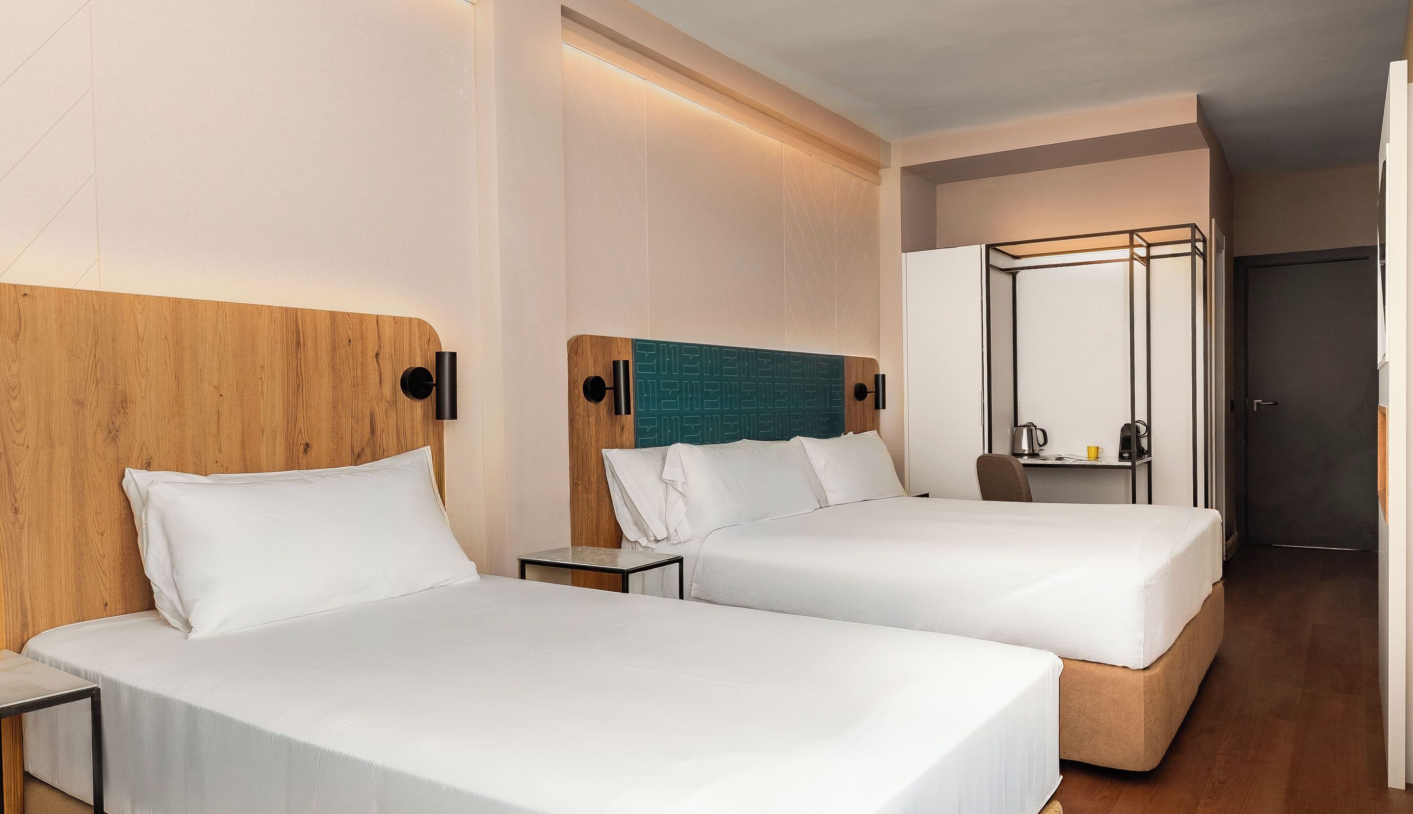 A hotel room with two beds, a wooden headboard, a mirror, and a small table with a kettle and cup.