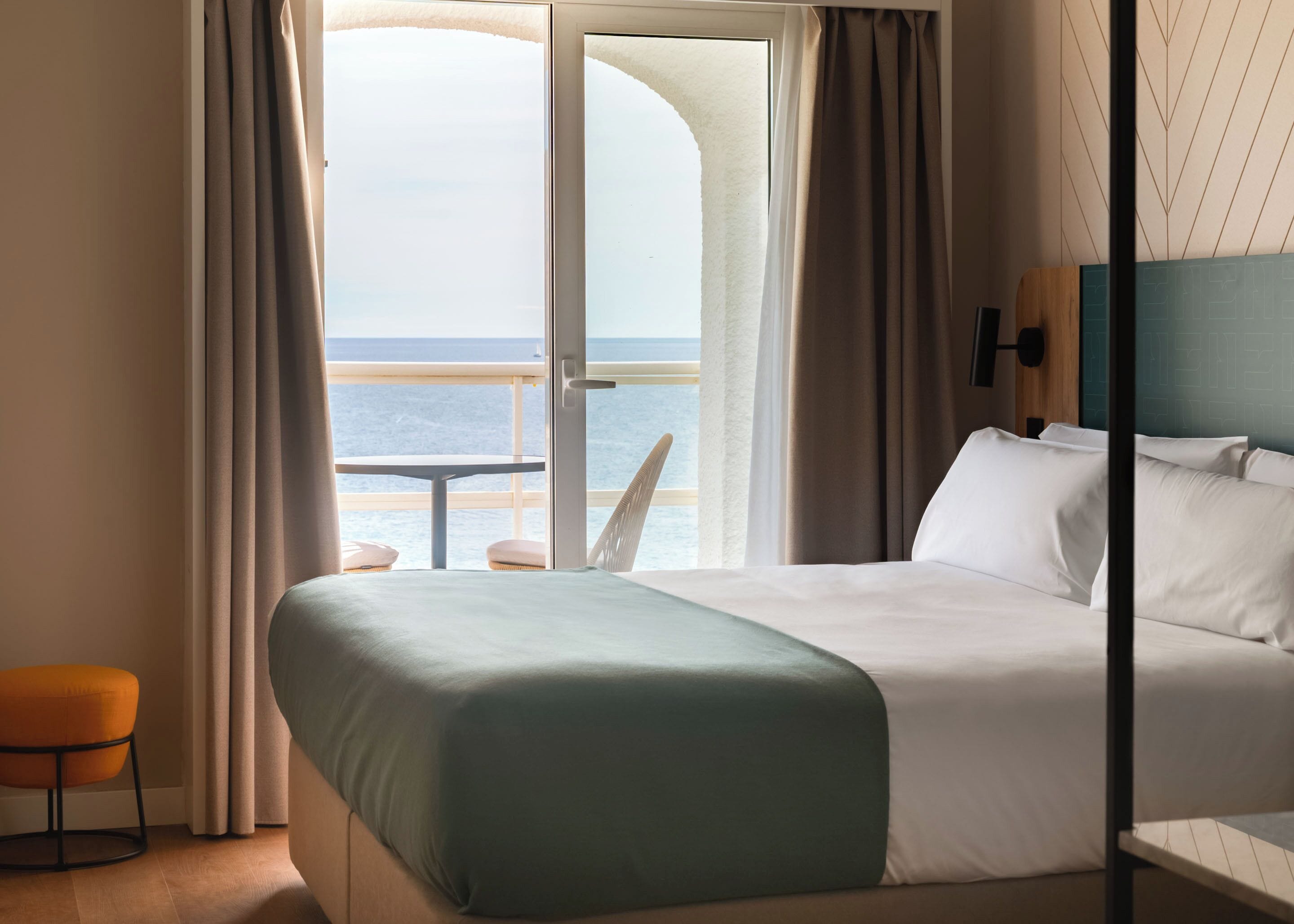 A hotel room with a bed, a small table, and a chair. There is a view of the ocean through large windows.