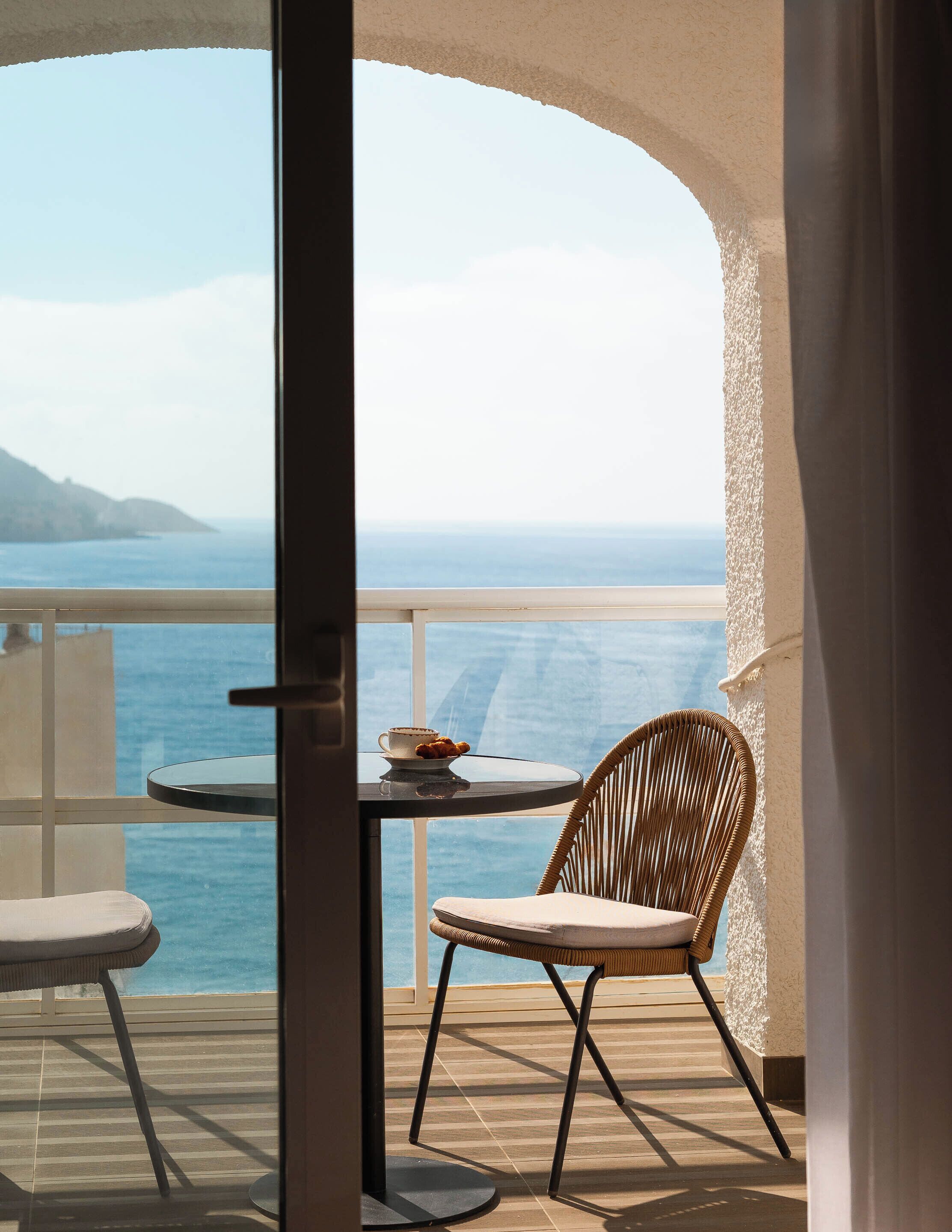 A balcony with a view of the sea, a table with a cup and pastries, and two wicker chairs.