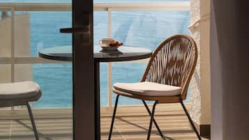 A balcony with a view of the sea, a table with a cup and pastries, and two wicker chairs.