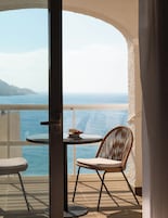 Un balcon avec vue sur la mer, une table avec une tasse et des pâtisseries, et deux chaises en osier.