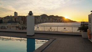 Una piscina en la azotea con vista a un paisaje urbano y montañas al atardecer.