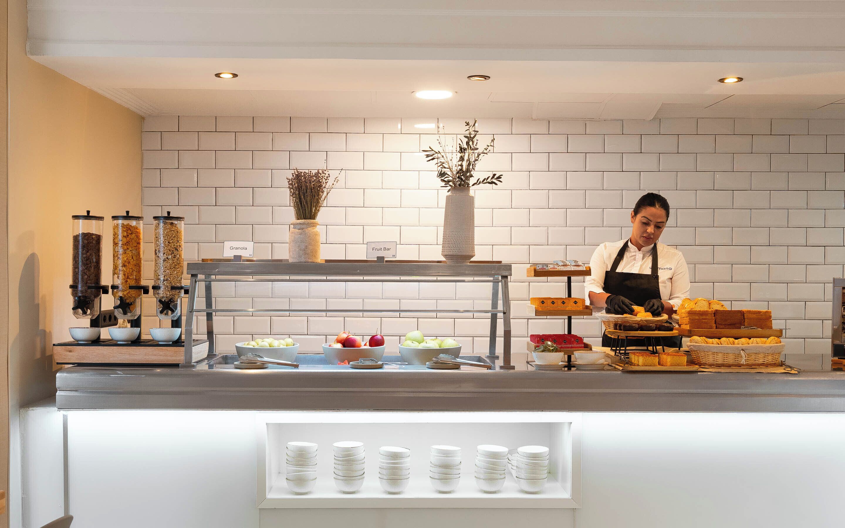 Una barra de desayuno con dispensador de café, una barra de frutas y una selección de panes y pasteles.